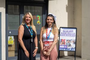 Two women stand smiling outside a work experience centre beside a sign reading Study With Us and Get Work Experience.