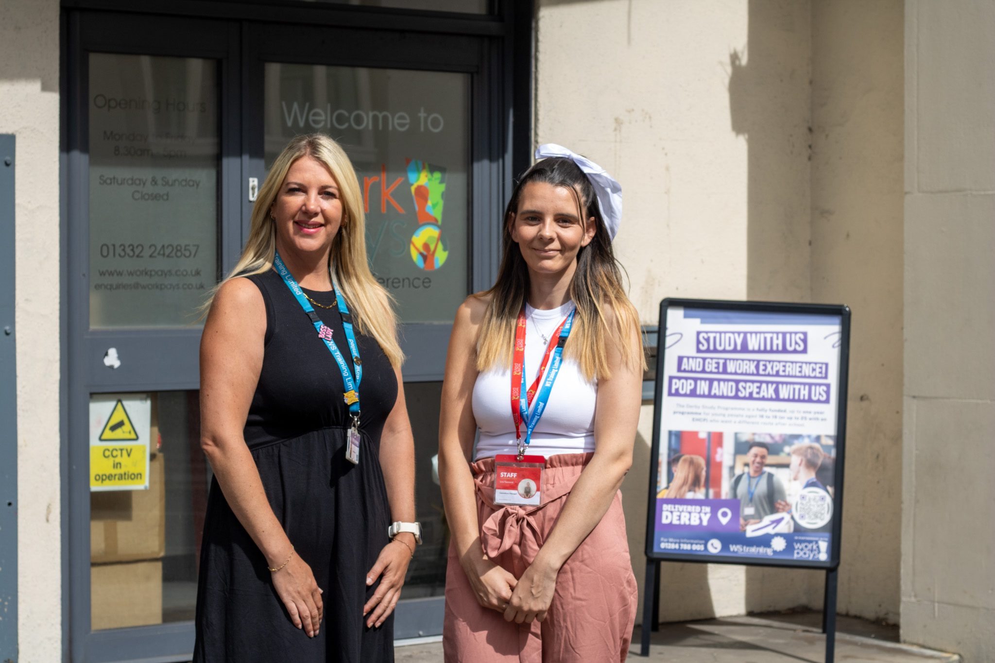 Two women stand smiling outside a work experience centre beside a sign reading Study With Us and Get Work Experience.